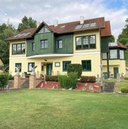 Ein großes zweistöckiges Haus mit einem braunen Dach und grüner Einfassung. Es verfügt über Solarpanels, einen Garten mit Pflanzen und eine Statue auf der Veranda.