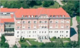 Ein Luftbild eines großen Gebäudes mit rotem Dach und vielen Fenstern. Es ist von einer großen Rasenfläche und Bäumen umgeben.