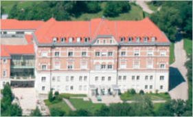 Ein Luftbild eines großen Gebäudes mit rotem Dach und vielen Fenstern. Es ist von einer großen Rasenfläche und Bäumen umgeben.