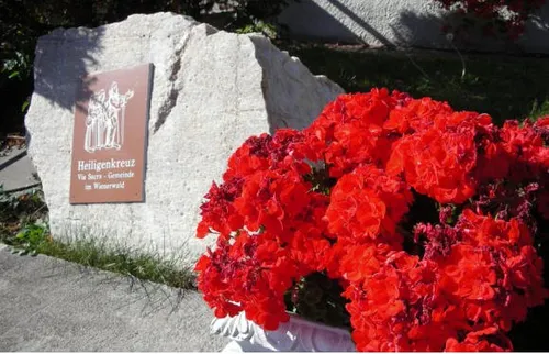 Eine Steinmauer mit einem Schild, das Heiligenkreuz, Via Sacra Gemeinde im Wienerwald besagt. Ein Ausschnitt roter Blumen in einem Topf.