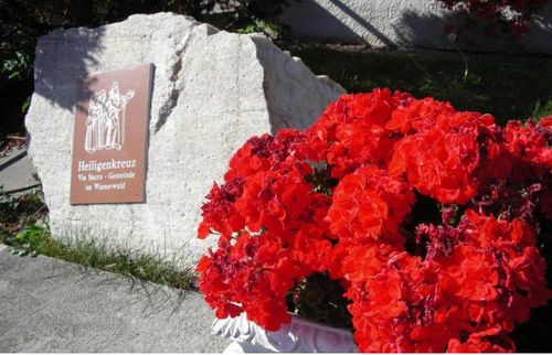 Eine Steinmauer mit einem Schild, das Heiligenkreuz, Via Sacra Gemeinde im Wienerwald besagt. Ein Ausschnitt roter Blumen in einem Topf.