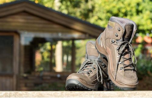 Ein Paar braune Wanderschuhe steht auf einer Betonfläche, mit einem hölzernen Gebäude im Hintergrund.