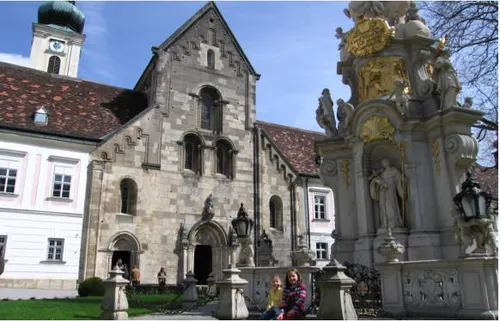Ein großes Steingebäude mit einem Glockenturm und Statuen steht vor einem blauen Himmel. Zwei junge Mädchen sitzen auf einem Zaun vor dem Gebäude.