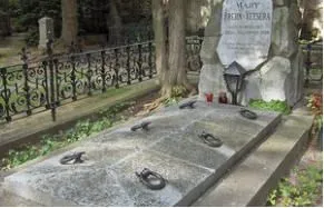 Ein Steindenkmal steht auf einem Friedhof mit einem schwarzen schmiedeeisernen Zaun und Bäumen. Es hat eine Plakette mit 'MAJTY FREDIN SETZERA' darauf. In der Nähe des Denkmals befinden sich auch einige Kerzen und eine Lampe.