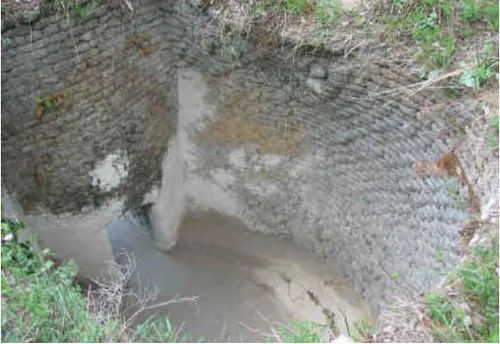 Ein Blick auf eine große, zylindrische Betonstruktur mit etwas Erosion und Wasser am Boden. Die Struktur ist von Grün umgeben.