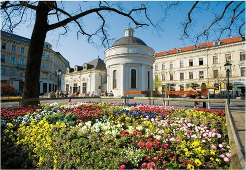 Ein Garten mit bunten Blumen vor einem historischen Gebäude mit einer Kuppel. Ein Paar sitzt auf einer Bank.