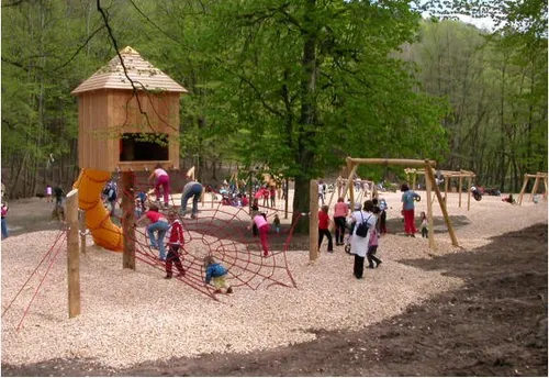 Kinder spielen in einem bewaldeten Bereich auf einem Spielplatz mit einer Holzkonstruktion, einer Rutsche und einem Kletternetz.