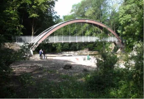Eine geschwungene Fußgängerbrücke aus Metall überspannt einen Fluss mit üppiger Grünanlage und Bäumen um sie herum. Zwei Personen stehen auf der Brücke.