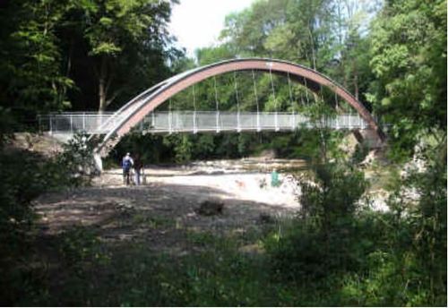 Eine geschwungene Fußgängerbrücke aus Metall überspannt einen Fluss mit üppiger Grünanlage und Bäumen um sie herum. Zwei Personen stehen auf der Brücke.