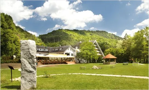 Ein landschaftliches Hotel mit Terrasse und Sitzgelegenheiten im Freien, umgeben von einem grünen Rasen und einem großen Felsen. Bäume und ein Berg sind im Hintergrund.