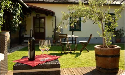 Eine Flasche Wein, zwei Weingläser und ein rotes Tuch stehen auf einem Tisch im Garten vor einem Haus, Bäumen und einem kleinen Tisch mit Stühlen.