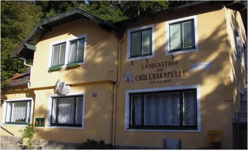 Die Vorderseite eines gelben Gebäudes mit Fenstern, beschriftet mit 'Landgasthof zur Cholerakapelle Fam. Safran'. Es gibt weiße Vorhänge in den Fenstern und ein dekoratives Huhn über einem der Fenster.