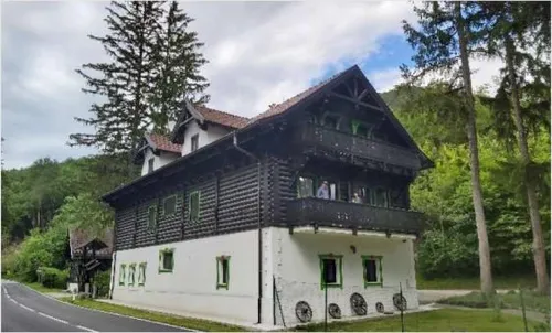 Ein großes Haus mit Balkon, umgeben von Bäumen und Gras. Das Haus hat ein braunes Dach und weiße Wände mit grünem Rand.