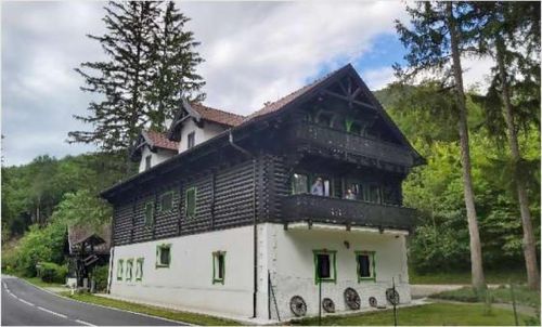 Ein großes Haus mit Balkon, umgeben von Bäumen und Gras. Das Haus hat ein braunes Dach und weiße Wände mit grünem Rand.