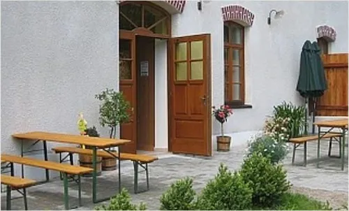 Ein Außenbereich mit Holztischen und Bänken in der Nähe eines Gebäudeeingangs mit offener Tür, umgeben von Pflanzen und Blumen, und einem geschlossenen Schirm.