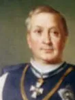 Ein Porträt eines älteren Mannes, der eine blaue Weste mit weißem Kragen und einer goldenen Kette mit Kreuzanhänger trägt. Er schaut mit einem leichten Lächeln nach vorne.