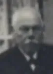 Ein altes Schwarz-Weiß-Foto zeigt einen älteren Mann mit einem Schnurrbart, der einen Anzug und eine Krawatte trägt.