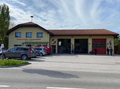 Ein Feuerwehrhaus namens Feuerwehr Siegenfeld mit geparkten Autos davor, stehenden Personen und einem Fahrrad in der Garage.