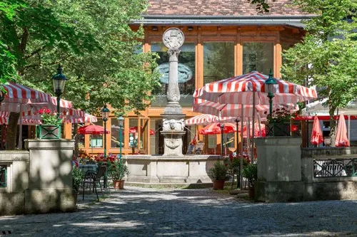Außensitzbereich mit Tischen und Stühlen unter roten und weißen Sonnenschirmen in der Nähe eines Steinbrunnens. Bäume und Straßenlaternen umgeben den Bereich.