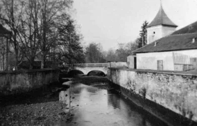 Ein Schwarz-Weiß-Foto zeigt einen Fluss, der durch eine Stadt fließt, mit einer Brücke und Gebäuden auf beiden Seiten. Bäume säumen die Flussufer.