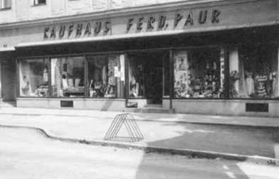 Ein altes Schwarz-Weiß-Foto zeigt ein Geschäft mit dem Schild 'KAUFHAUS FERD. PAUR'. Das Geschäft hat Glasfenster, die verschiedene Artikel präsentieren.
