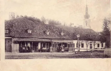 Ein altes Foto zeigt ein Gebäude mit Menschen, die davor stehen. Es hat ein steiles Dach und mehrere Fenster. Im Hintergrund sind Bäume und ein Kirchturm zu sehen.