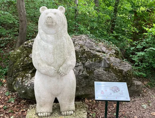 Eine große Steinbärenstatue steht auf einem Felsen im Wald. Sie hat ihre Hände auf dem Bauch. Dahinter befindet sich ein Informationsschild mit Informationen über Bären.