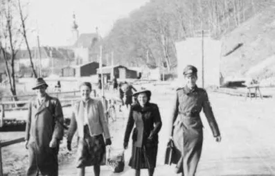 Ein Schwarz-Weiß-Foto zeigt vier Personen, die auf einer Straße gehen. Zwei Frauen tragen Taschen. Ein Mann trägt eine Militäruniform. Sie gehen an Gebäuden und Bäumen vorbei.