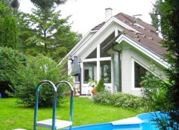 Ein weißes Haus mit braunem Dach und Glasfenstern steht in einem Garten mit grünem Rasen, Büschen und einem blauen Schwimmbecken mit Sprungbrett.