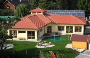 Eine Luftaufnahme eines gelben Hauses mit rotem Dach und Solarmodulen. Das Haus hat einen Garten, einen grünen Rasen und einen kleinen Swimmingpool. In der Nähe stehen einige Bäume und andere Häuser.