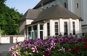 Ein weißes Gebäude mit einem braunen Dach und mehreren bogenförmigen Fenstern steht vor einem Garten mit bunten Blumen. Bäume und eine Statue sind im Hintergrund zu sehen.