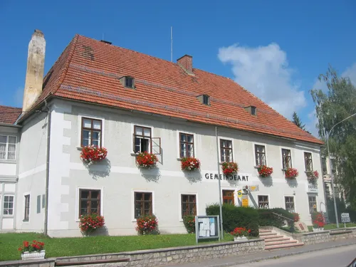 Ein weißes Gebäude mit einem roten Dach, mehreren Fenstern, die mit roten Blumenkästen geschmückt sind, und einem Schild mit der Aufschrift 'Gemeindeamt'. Das Gebäude ist von grünem Gras und Büschen umgeben, mit einer Treppe, die zum Eingang führt.