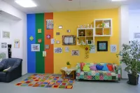 Ein farbenfroher Raum mit bunten Wänden, einem gemusterten Teppich und einem gemütlichen Sofa, das mit bunten Kissen geschmückt ist. Verschiedene gerahmte Bilder und Kunstwerke schmücken die Wände. Ein Tisch mit einem Topf steht neben dem Sofa.