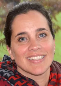 Eine Frau mit dunklem Haar und blauen Augen lächelt und trägt einen Schal. Sie hat einen Muttermal auf ihrer rechten Wange.