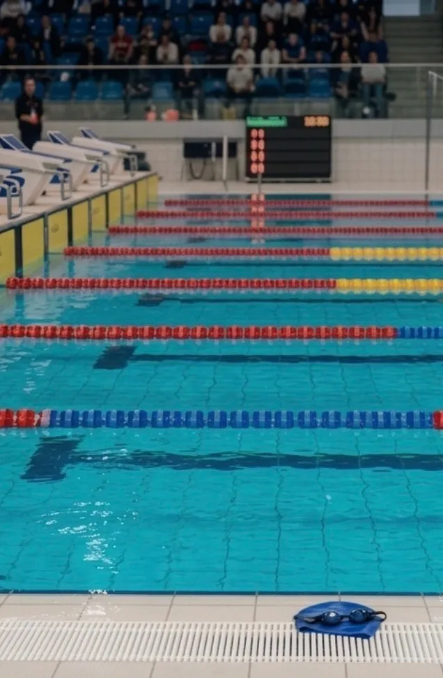 Abschnittsbild: Ein leerer Schwimmpool mit durch rote und blaue Schwimmer markierten Bahnen. Ein Anzeigetafel ist über dem Pool sichtbar, und eine Person steht in der Nähe der Poolkante.
