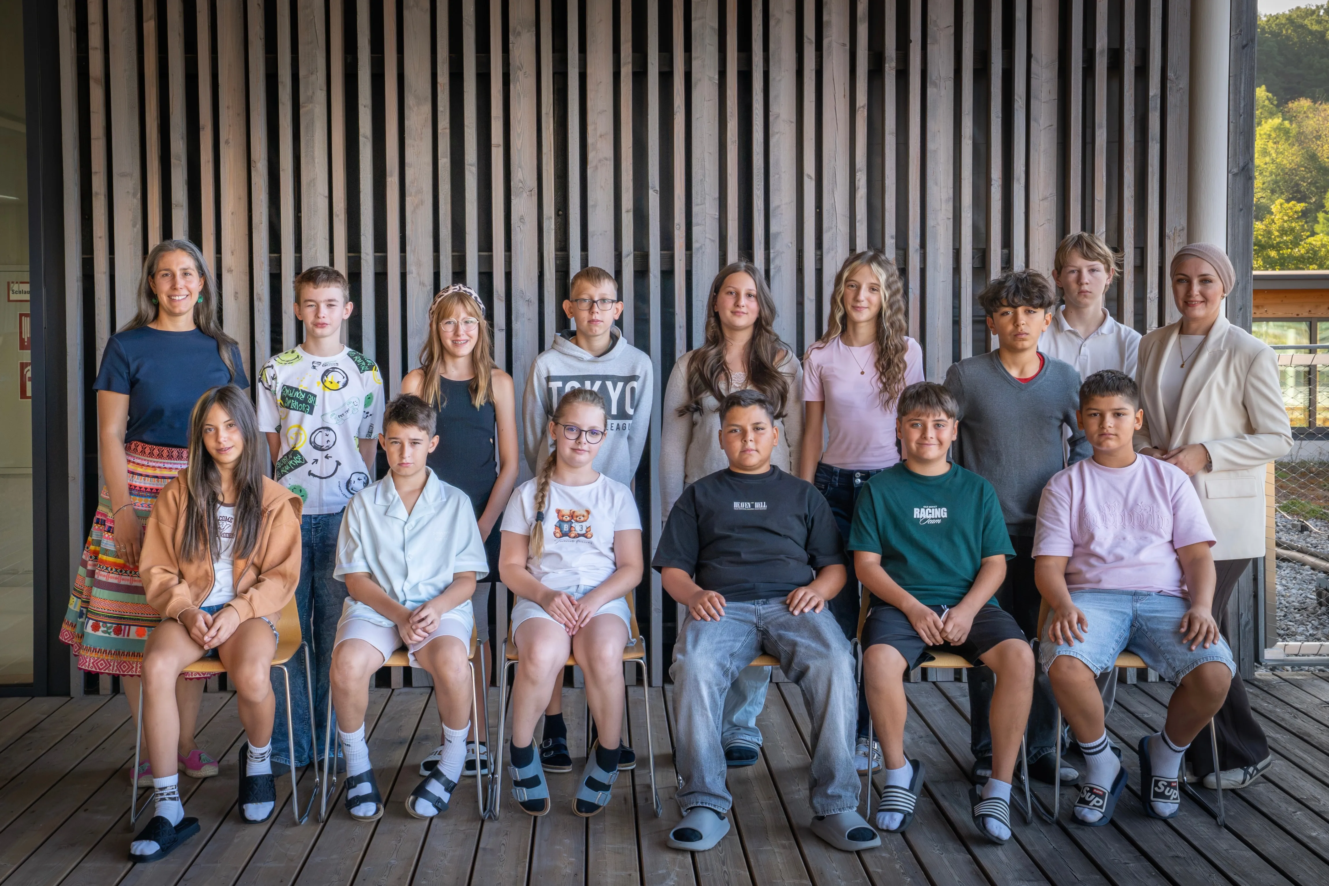 Eine Gruppe junger Menschen, sowohl Jungen als auch Mädchen, sitzen auf Stühlen vor einer Holzwand. Sie alle lächeln und schauen in die Kamera.