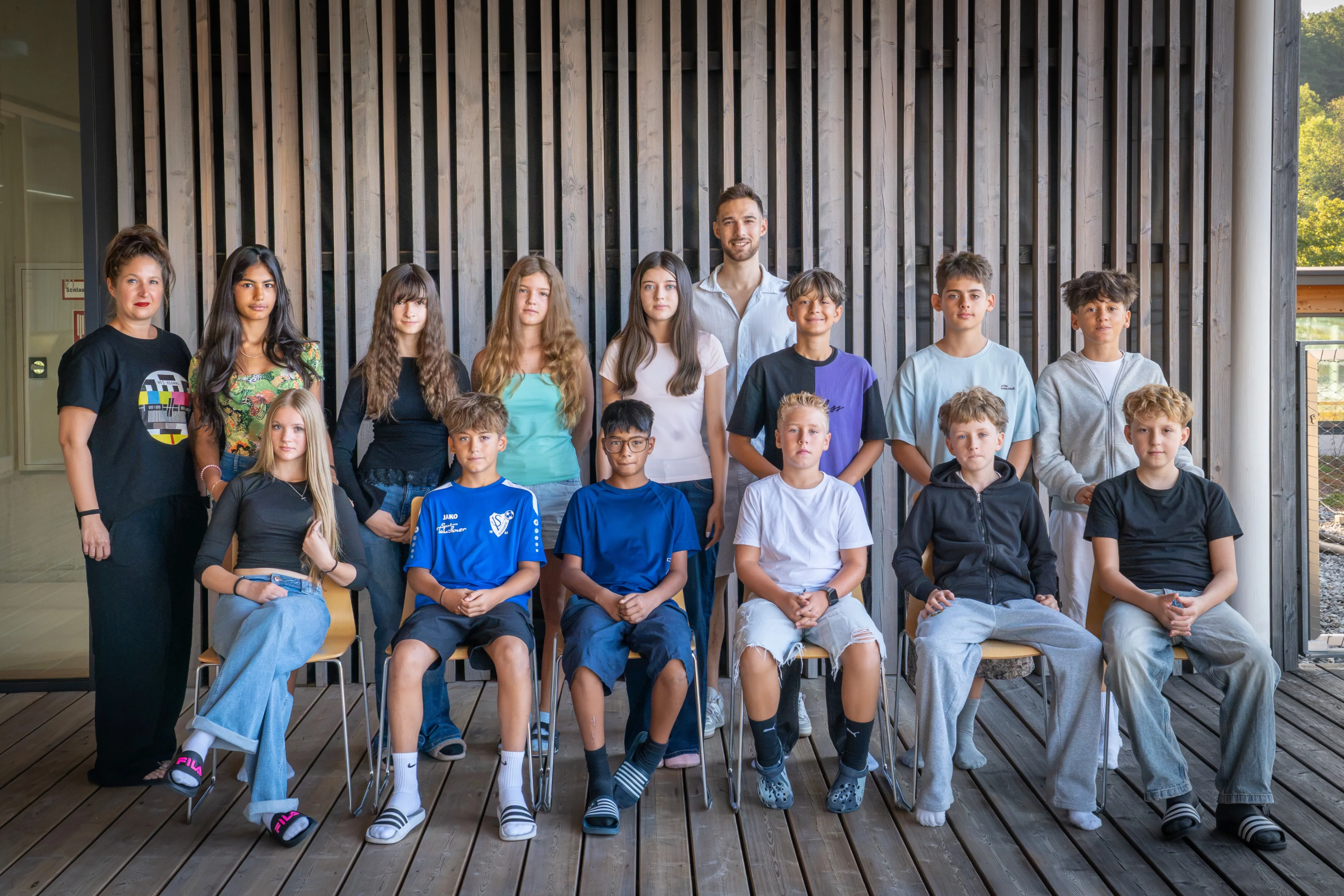 Eine Gruppe junger Menschen, darunter Jungen und Mädchen, sitzen auf Stühlen vor einer Holzwand. Sie alle lächeln und posieren für ein Foto.