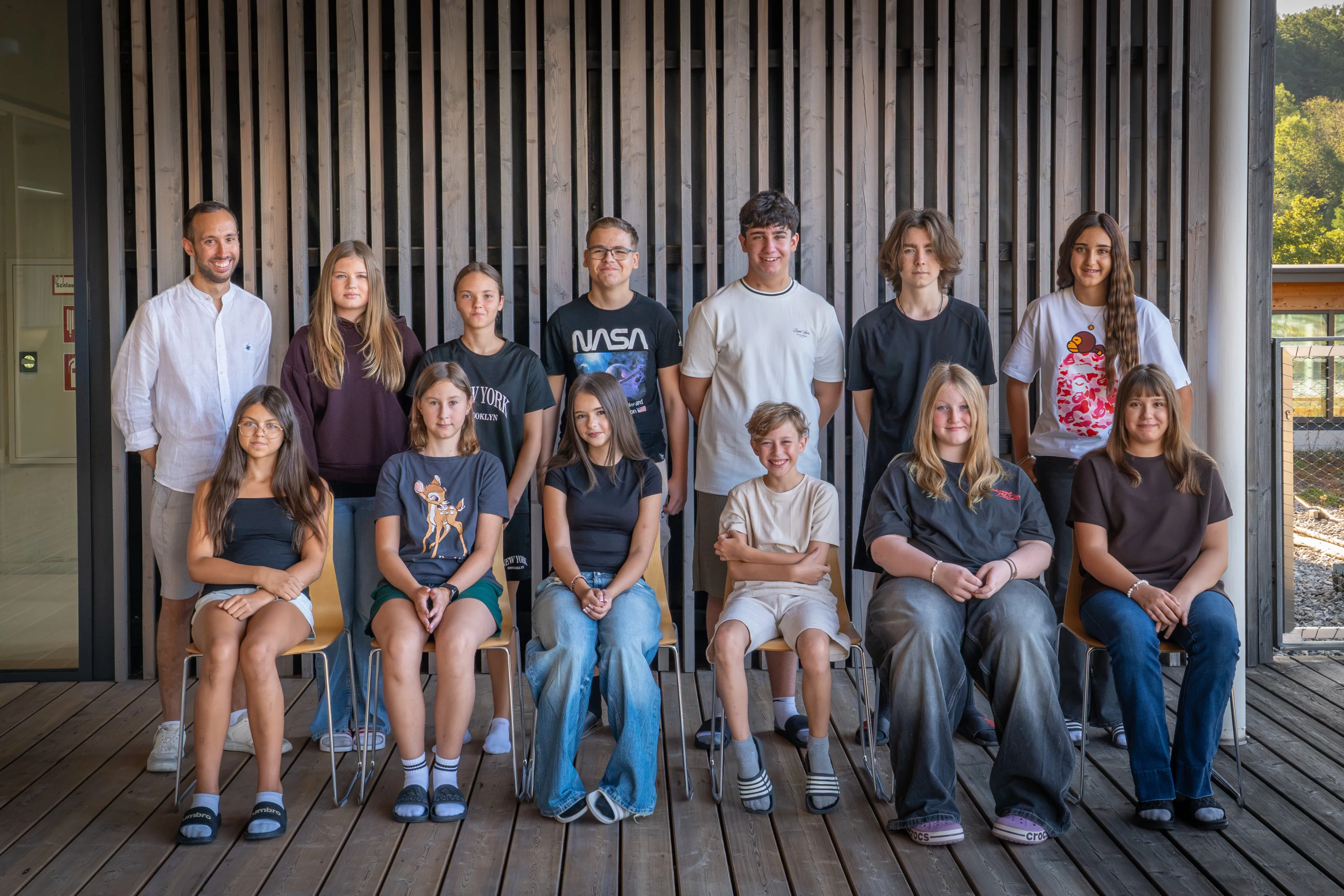 Eine Gruppe von Teenagern und jungen Erwachsenen posiert für ein Foto auf einer Holzterrasse. Sie tragen Freizeitkleidung und einige haben eine Brille. Die Holzwand hinter ihnen hat vertikale Streifen.