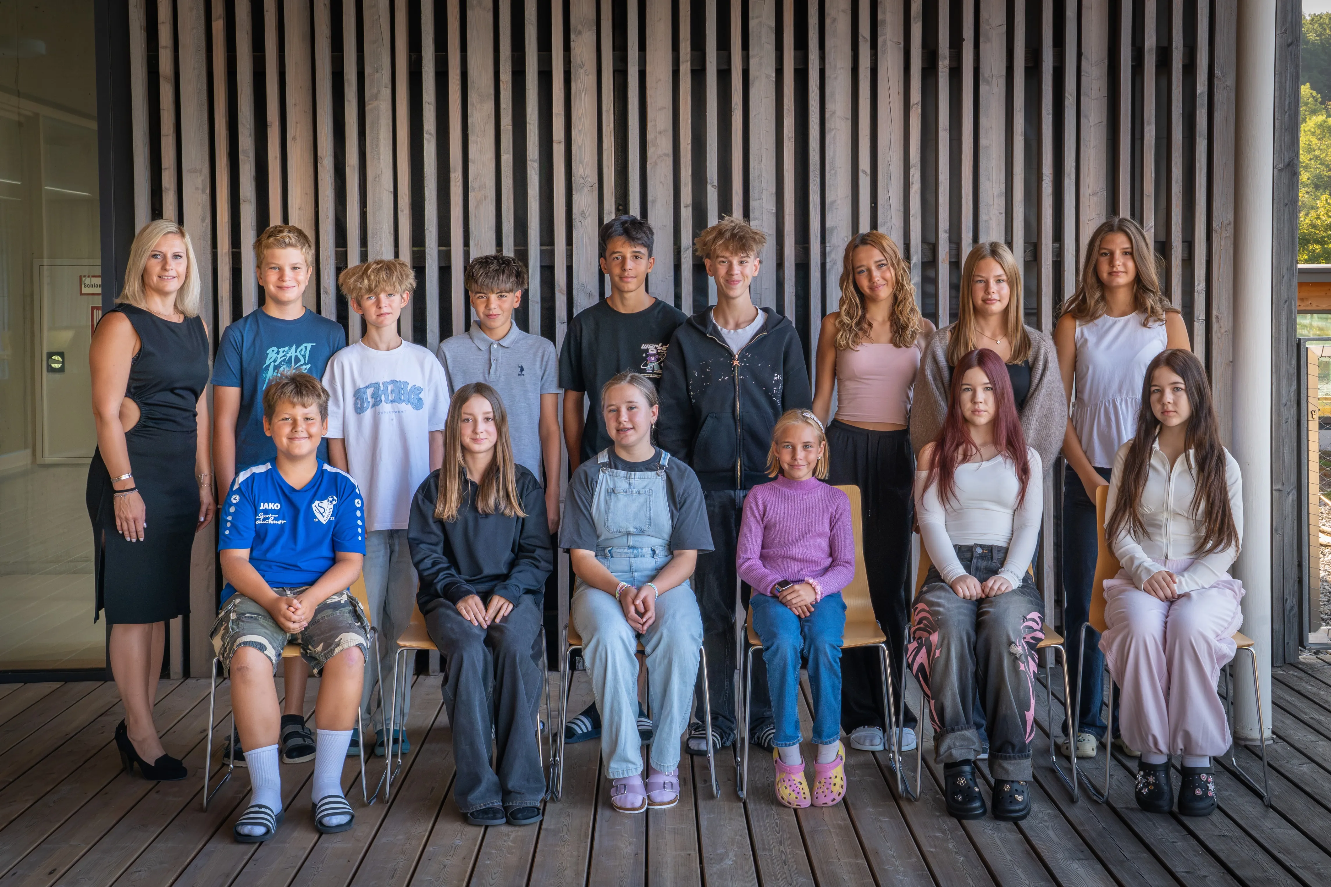Eine Gruppe von Teenagern und jungen Erwachsenen posiert für ein Foto vor einer Holzwand. Sie sitzen und stehen in verschiedenen Posen.