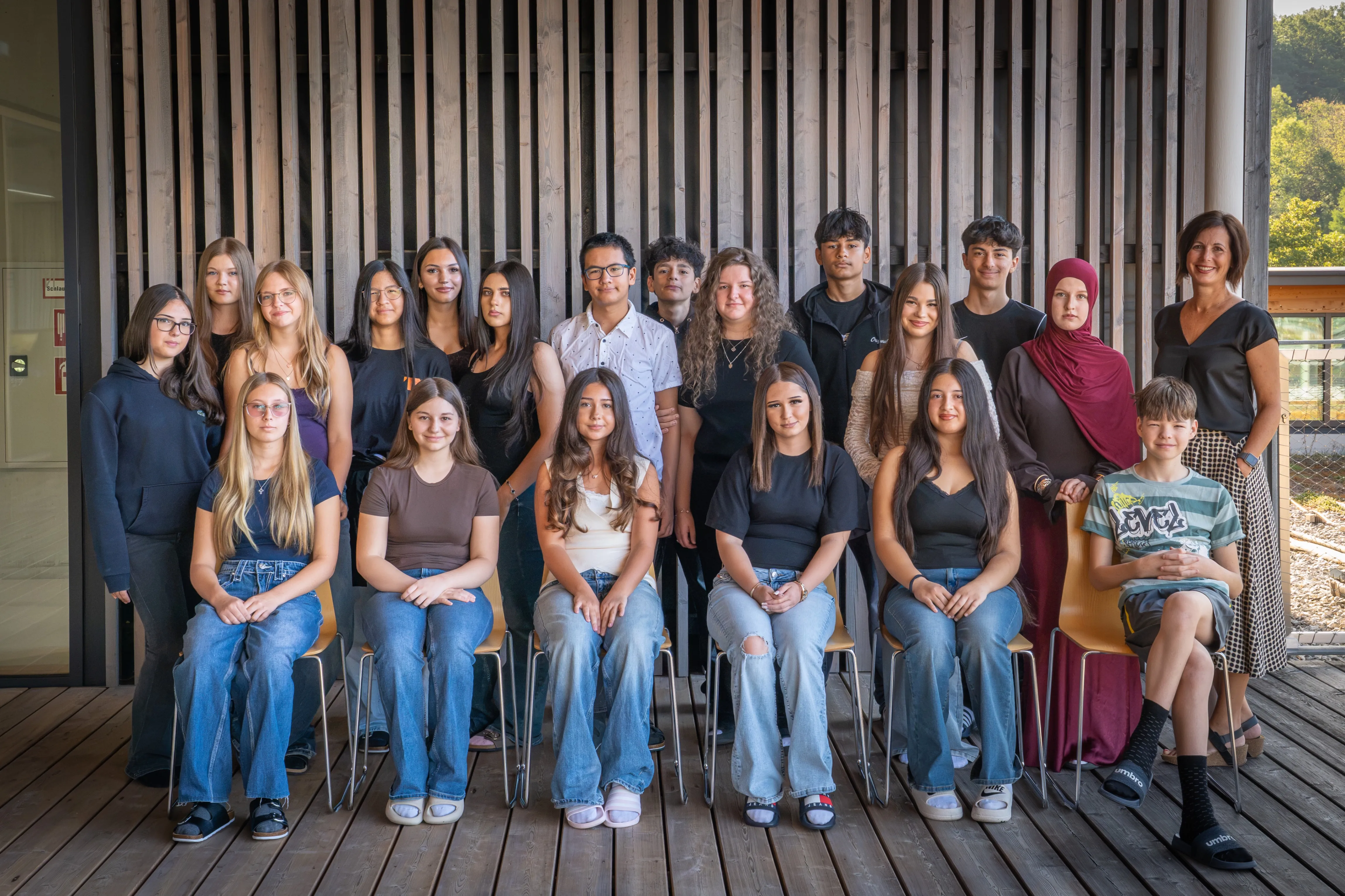 Eine Gruppe junger Menschen, sowohl Männer als auch Frauen, sitzen auf Stühlen vor einer Holzwand. Sie alle lächeln und scheinen für ein Gruppenfoto zu posieren.