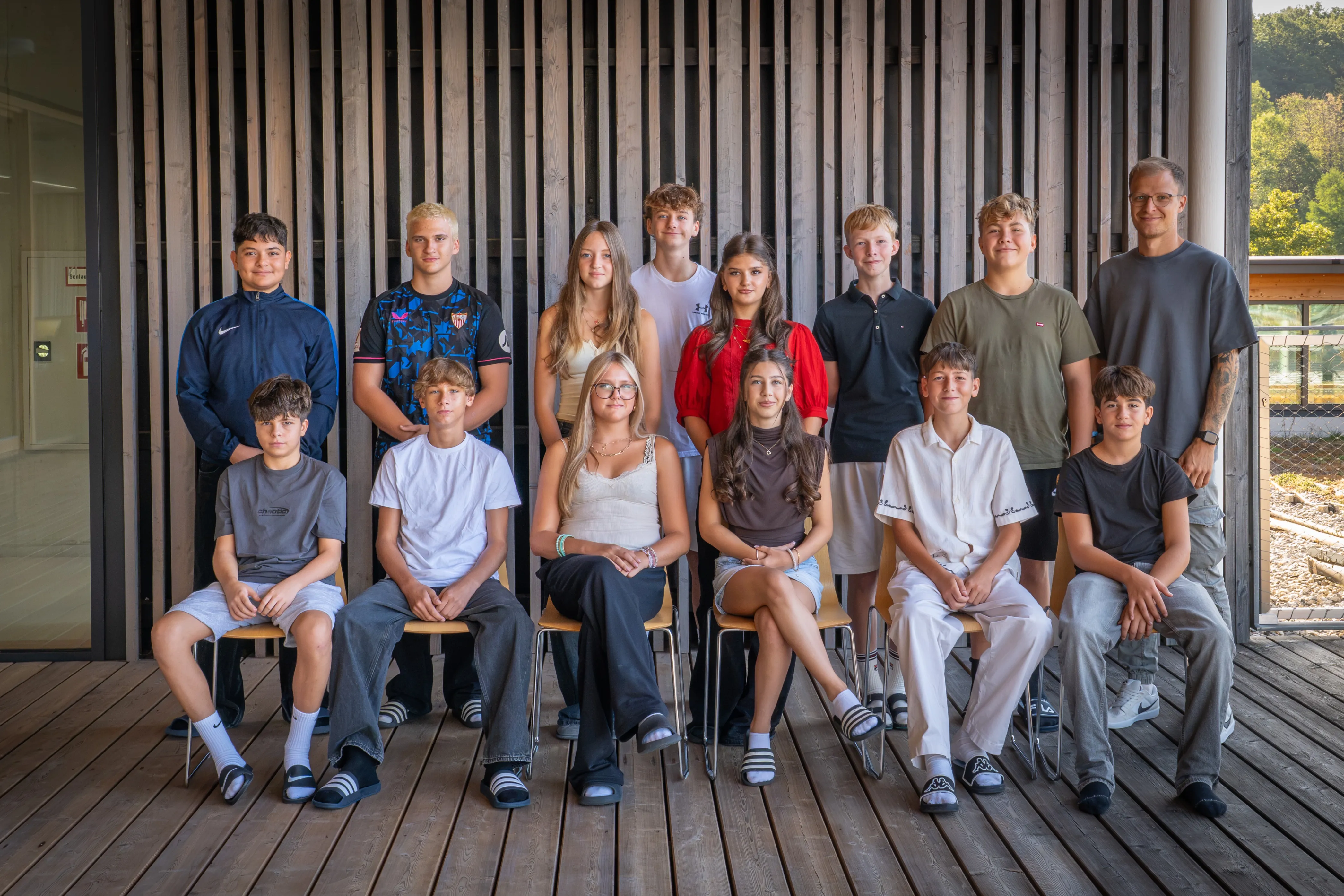 Eine Gruppe junger Menschen, sowohl Jungen als auch Mädchen, sitzen in einer Reihe auf Stühlen, wahrscheinlich für ein Gruppenfoto. Sie sind alle lässig gekleidet.