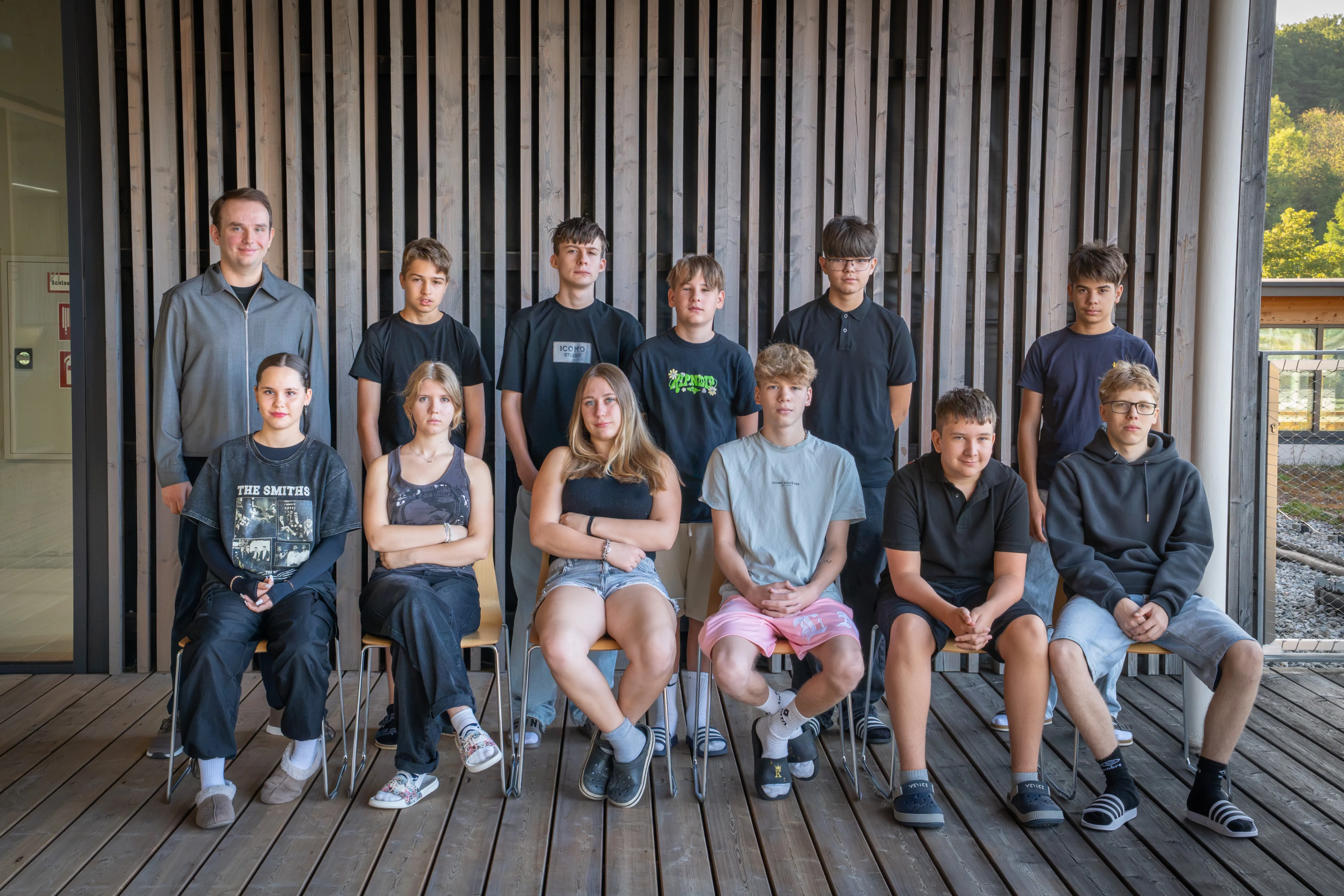Eine Gruppe junger Menschen, sowohl Jungen als auch Mädchen, posiert für ein Foto vor einer Holzwand. Sie sind alle lässig gekleidet.