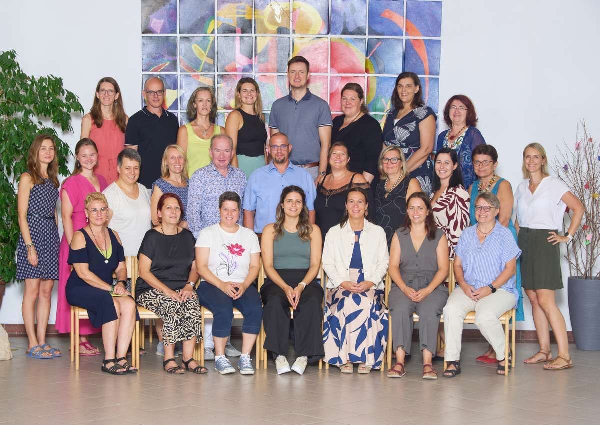 Eine Gruppe von Menschen, sowohl Männer als auch Frauen, posiert für ein Foto vor einer farbenfrohen Wandmalerei. Sie alle lächeln und einige tragen Brillen.