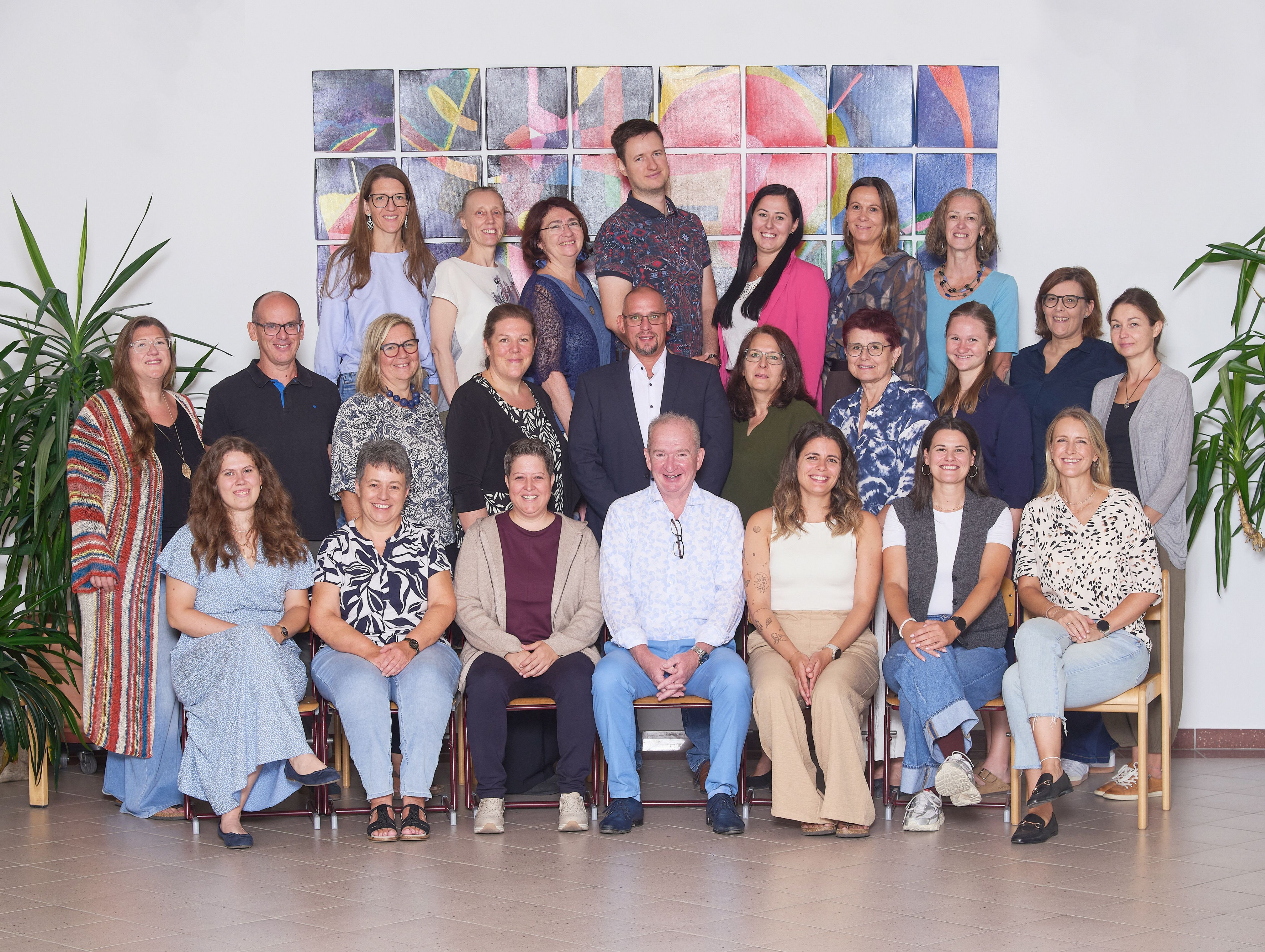 Eine Gruppe von Menschen, sowohl Männer als auch Frauen, posiert für ein Foto vor einer farbenfrohen Wand. Sie alle lächeln und scheinen Teil eines Teams oder einer Organisation zu sein.