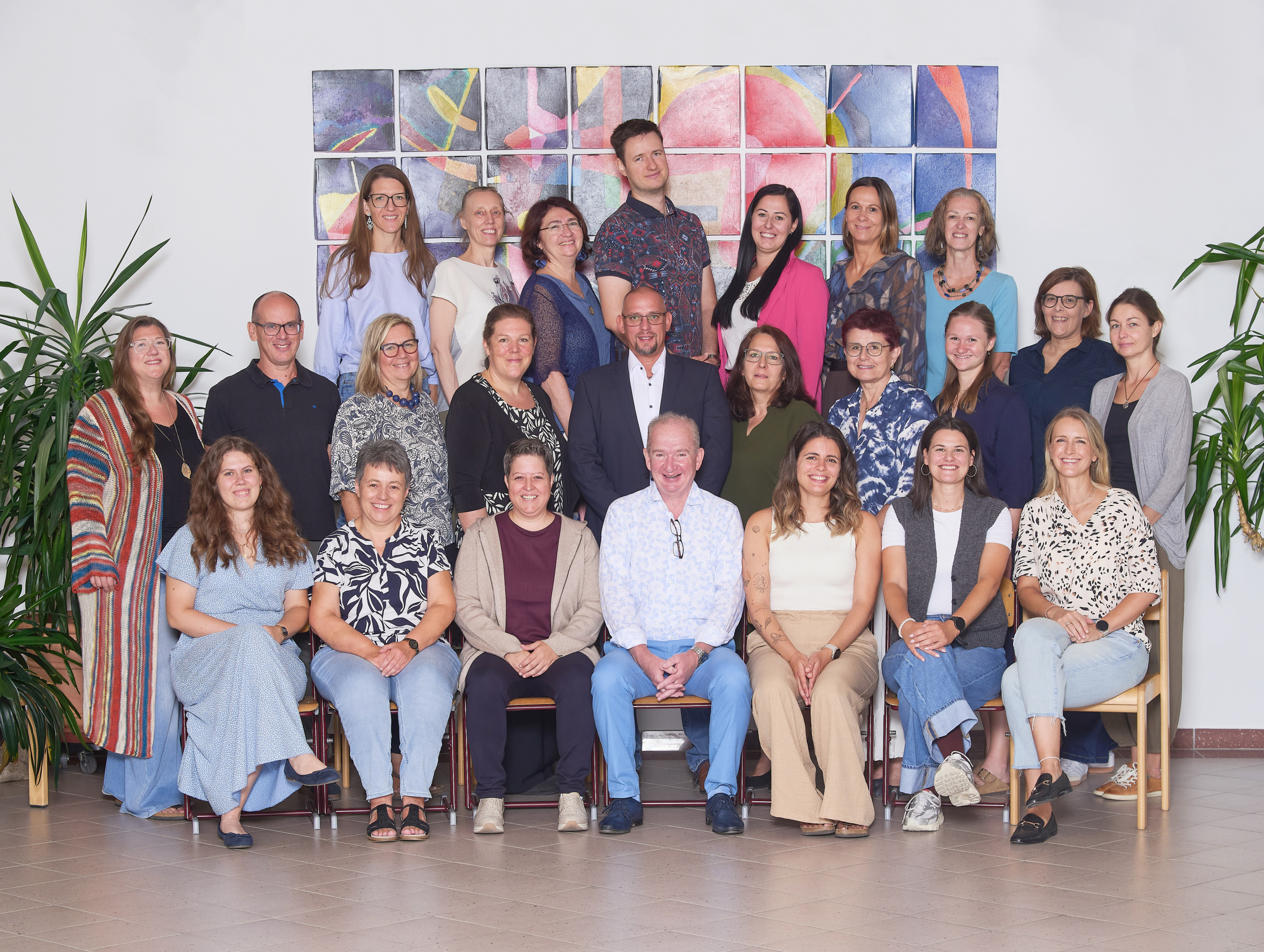 Eine Gruppe von Menschen, sowohl Männer als auch Frauen, posiert für ein Foto vor einer farbenfrohen Wand. Sie alle lächeln und scheinen Teil eines Teams oder einer Organisation zu sein.