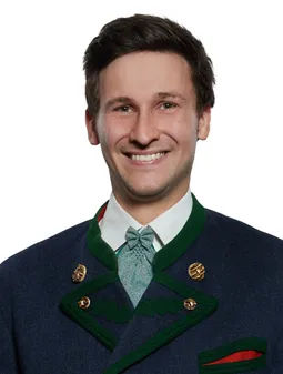 Ein lächelnder Mann in einem dunkelblauen Anzug mit grünem Kragen, weißem Hemd und blauer Krawatte posiert für ein Foto.