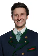 Ein Mann in traditioneller bayerischer Kleidung mit lächelndem Gesicht, trägt eine grün-blaue Jacke mit goldenen Knöpfen am Kragen.