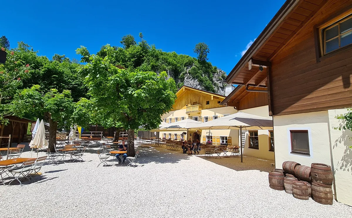Sitzbereich im Freien eines Restaurants. Tische und Stühle sind unter Schirmen angeordnet. Ein Mann sitzt allein und eine Gruppe sitzt an einem anderen Tisch. Das Gebäude ist holzfarben, mit gelben und weißen Wänden. Bäume und ein Berg sind im Hintergrund.