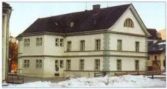 Ein großes weißes Gebäude mit schwarzem Dach steht in einer verschneiten Landschaft, umgeben von Schnee. Es hat mehrere Fenster und einen Schornstein auf dem Dach. Vor dem Gebäude befindet sich ein Zaun.