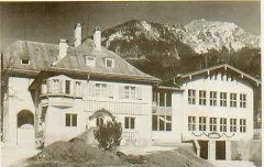 Ein altes Schwarz-Weiß-Foto zeigt ein großes Gebäude mit einem geneigten Dach, mehreren Schornsteinen und vielen Fenstern. Im Hintergrund ist eine verschneite Bergkette zu sehen.